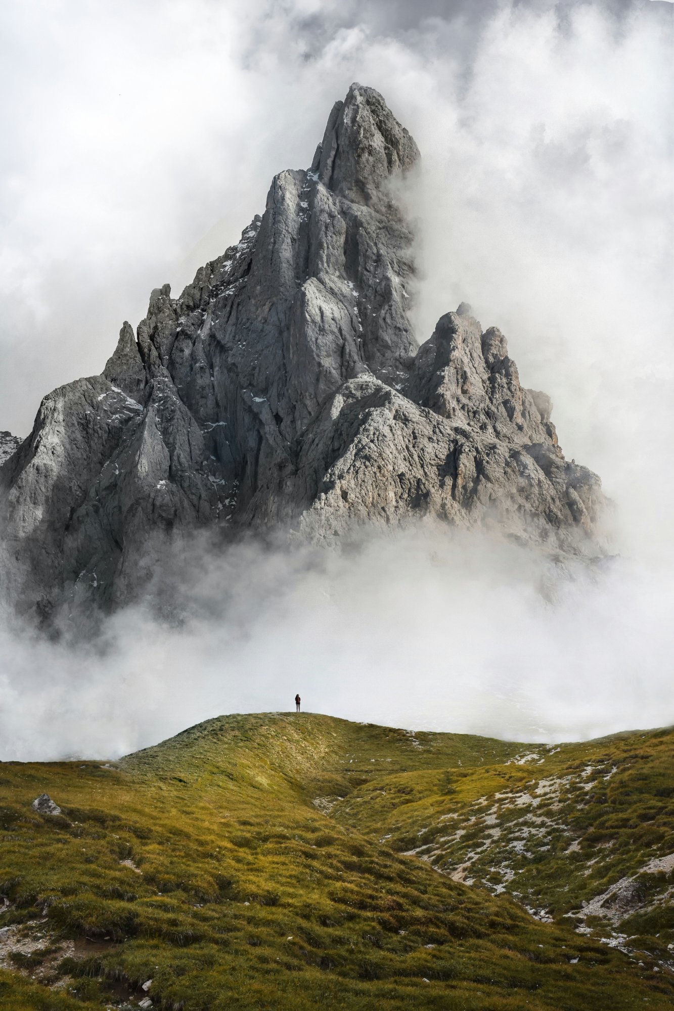 A person standing on a green hill with a mountain emerging from clouds, grounded in nature