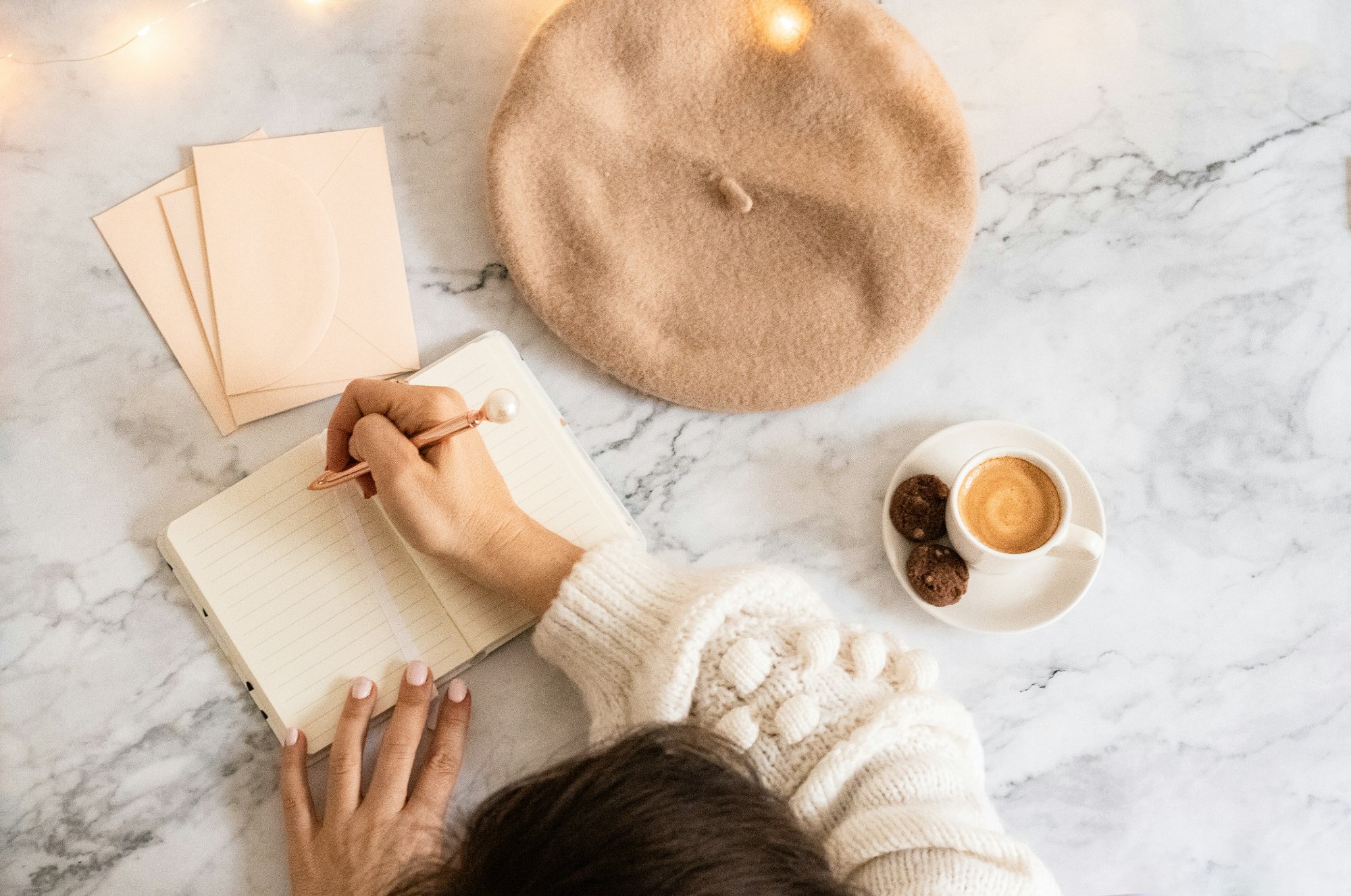 Overhead view of a woman writing in a notebook at a marble desk with coffee and letters