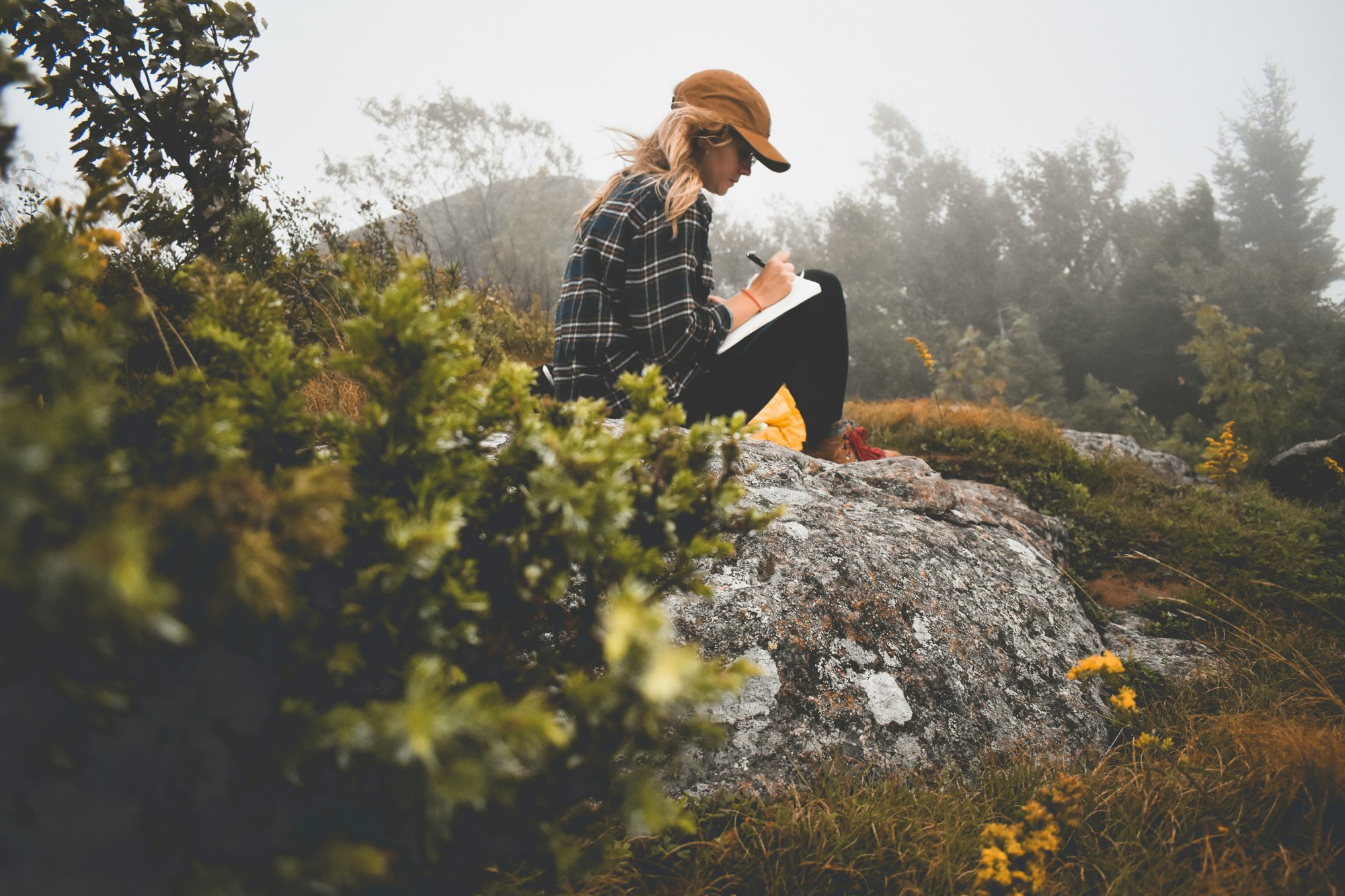 Woman sitting on a rock in misty nature, journaling in a notebook to process anxiety