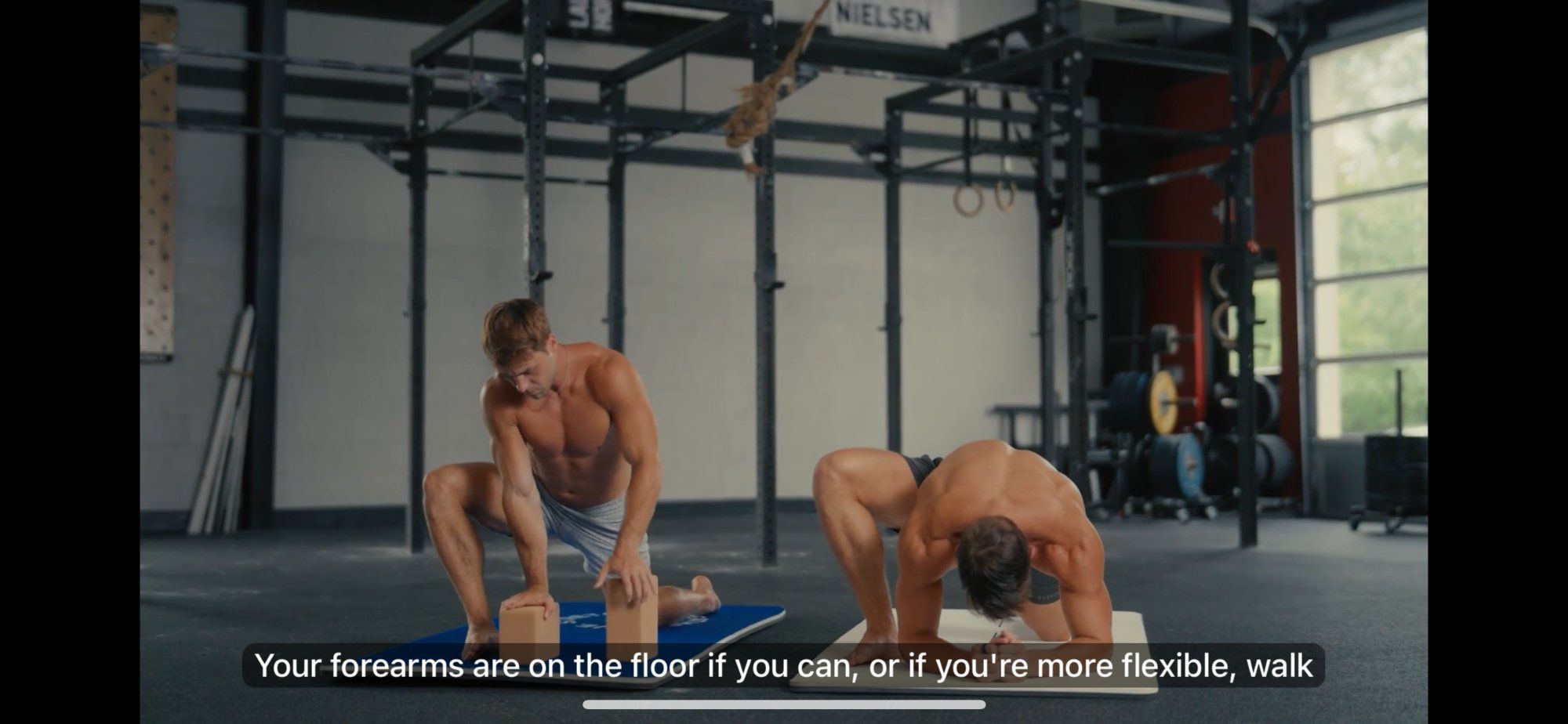 A Pliability session in progress: two athletes in a gym demonstrating a stretch, with voice guidance subtitles showing 'Your forearms are on the floor if you can, or if you're more flexible, walk'